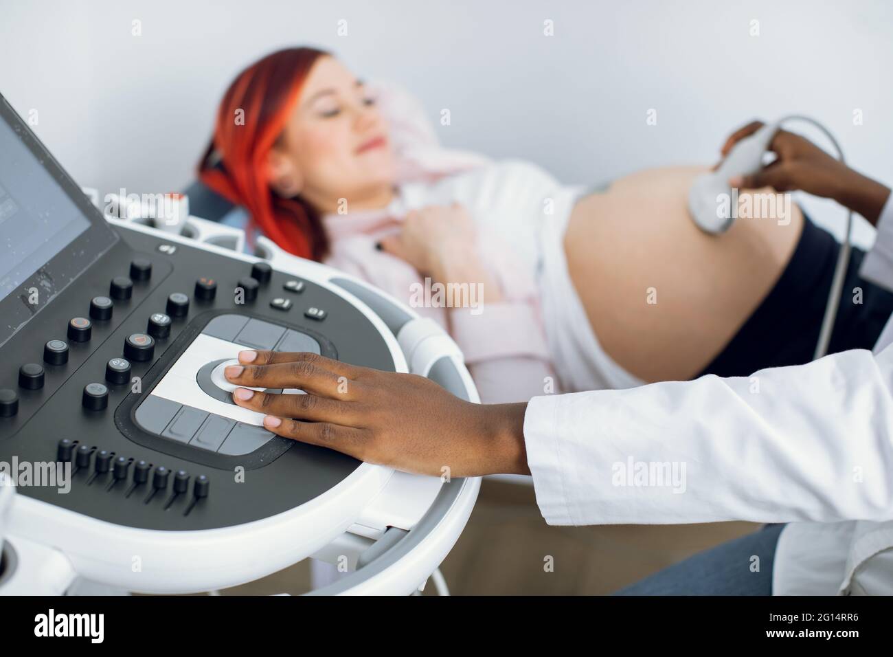 Primo piano immagine ritagliata di ostetrica afro-medica femminile, premendo i pulsanti su un pannello di controllo che esegue la procedura ecografica per la donna incinta con capelli rossi luminosi Foto Stock