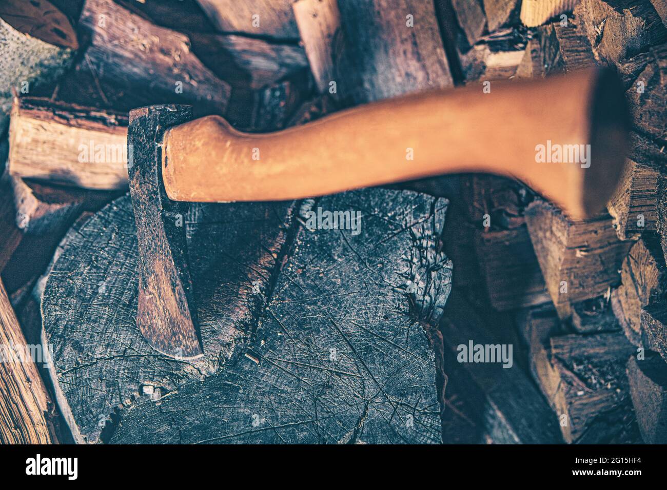 Capannone di legno con un'ascia minacciosa e un grande tronco con un sacco di legna da ardere impilata sullo sfondo. Leggera incidenza nel primo piano del bosco. Foto Stock