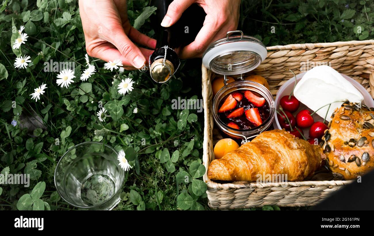 Cestino rustico da picnic con croissant, marmellata, formaggio, pane su prato verde e sfondo a margherita. Le mani femminili aprono una bottiglia di sidro bevanda. Foto Stock