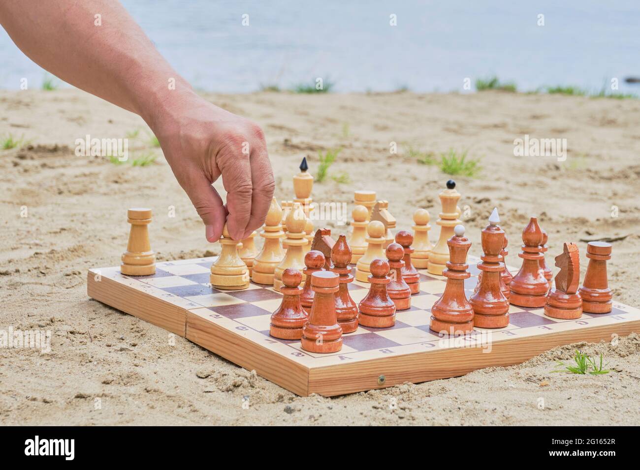 La mano di un uomo anziano che tiene una pedina per una mossa in un gioco di scacchi sullo sfondo di una spiaggia sabbiosa vicino al lago. Concetto di disintossicazione digitale Foto Stock