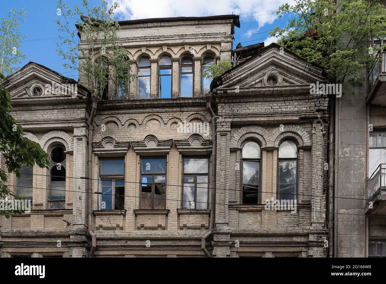 La facciata di una vecchia casa sbriciolare con vetri rotti nelle finestre e alberi germogliati sul tetto. Casa infestata, vecchia casa spaventosa, architettura rec Foto Stock
