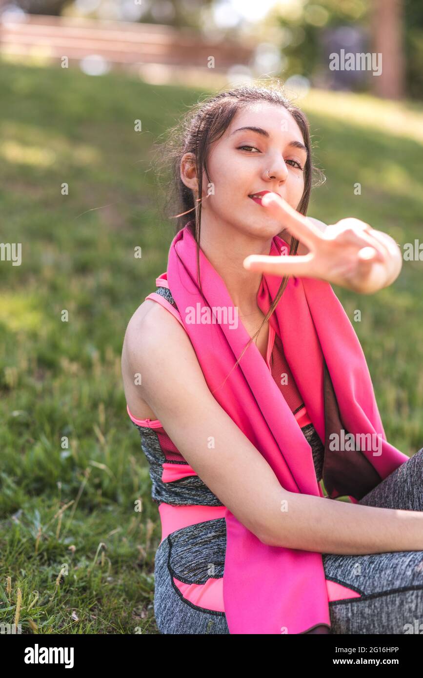 ritratto verticale di una giovane donna che fa il gesto della vittoria alla macchina fotografica. Si sta vestendo abiti sportivi e si siede sull'erba Foto Stock