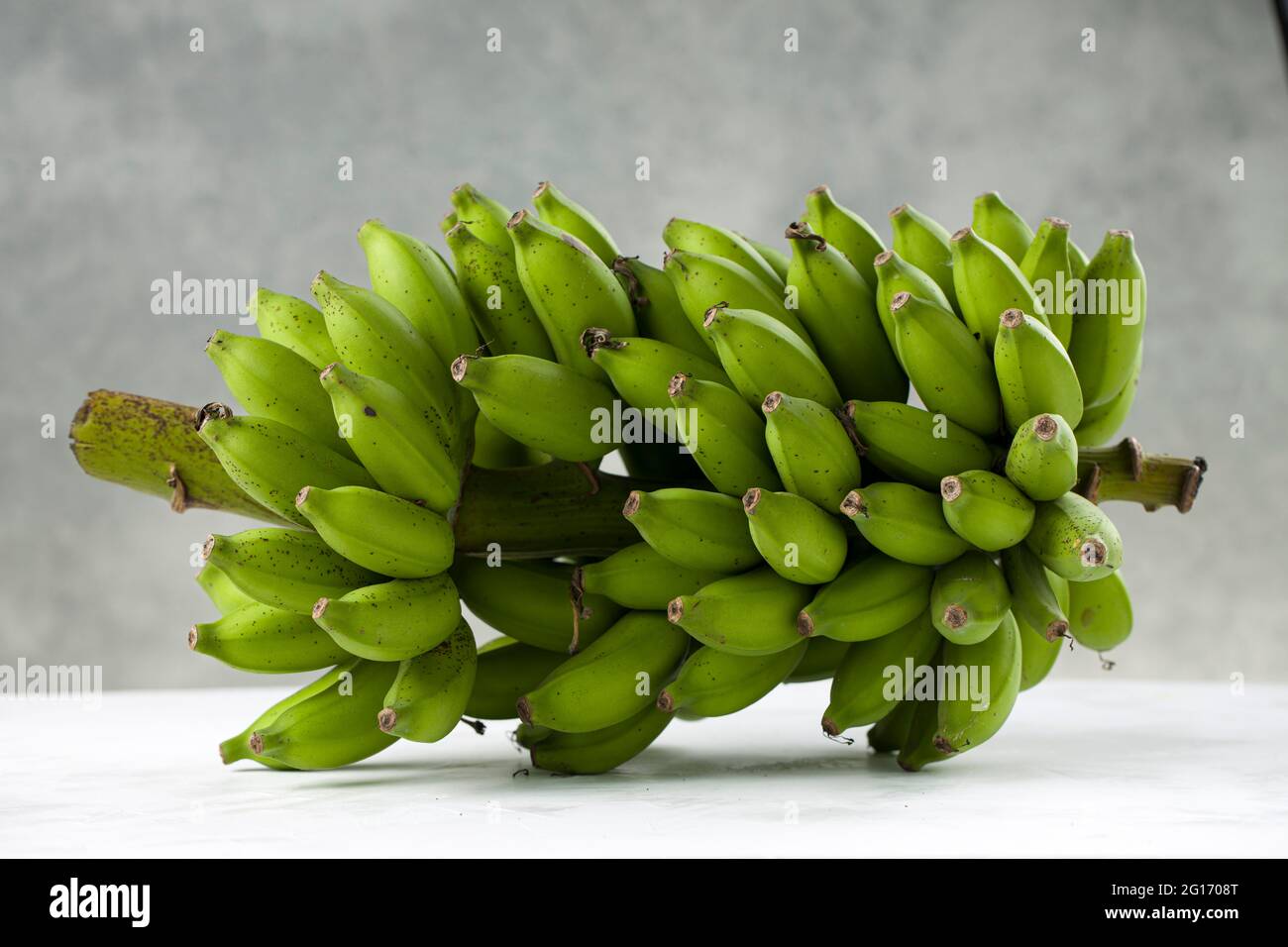 Mazzetto di banana grezza o di banana verde posto su una superficie bianca con fondo grigio testurizzato, isolato. Foto Stock