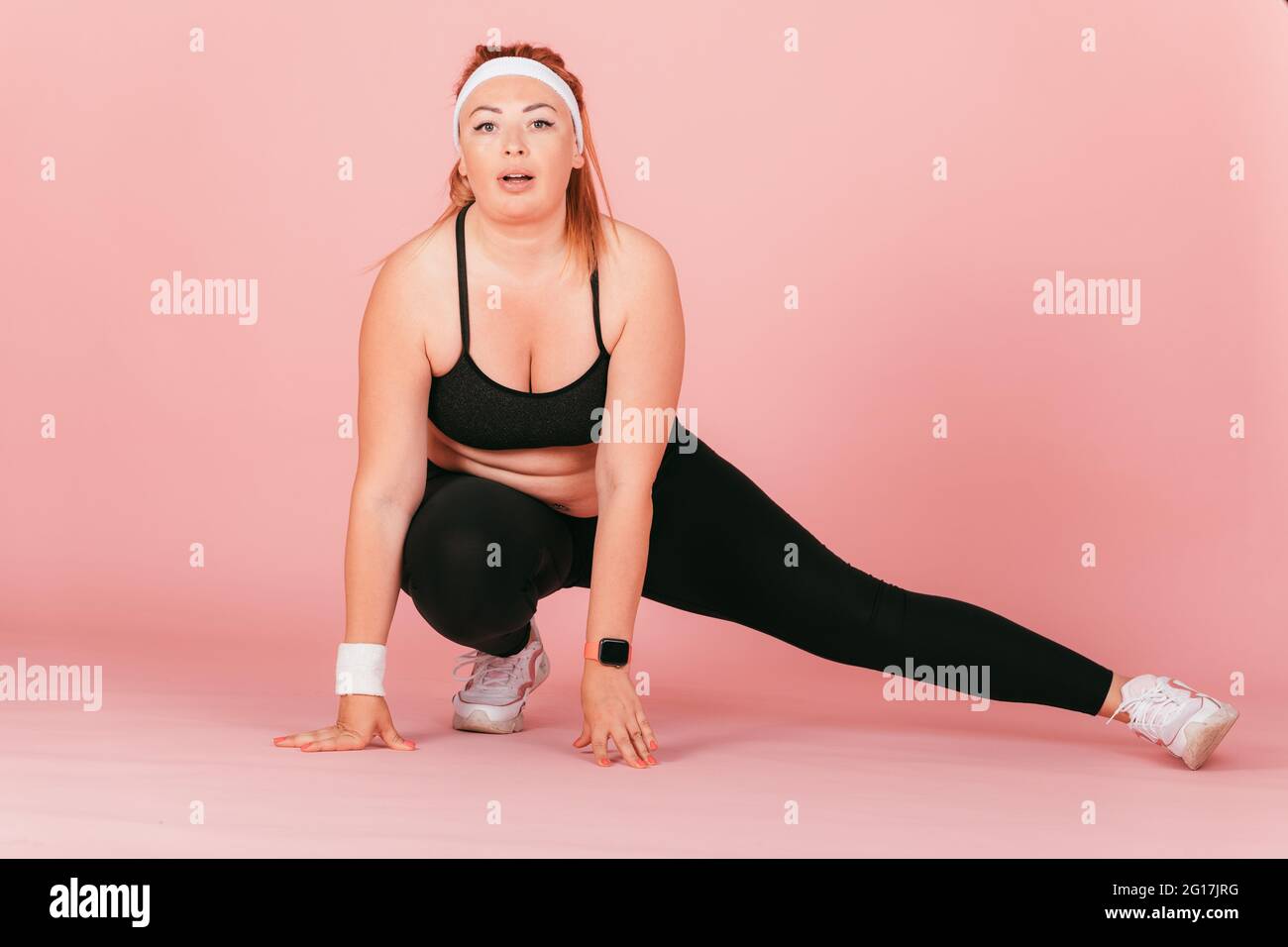 Ritratto a lunghezza intera della donna in groppa che fa esercizio fisico, allungando le gambe in squat, sfondo rosa. Foto Stock