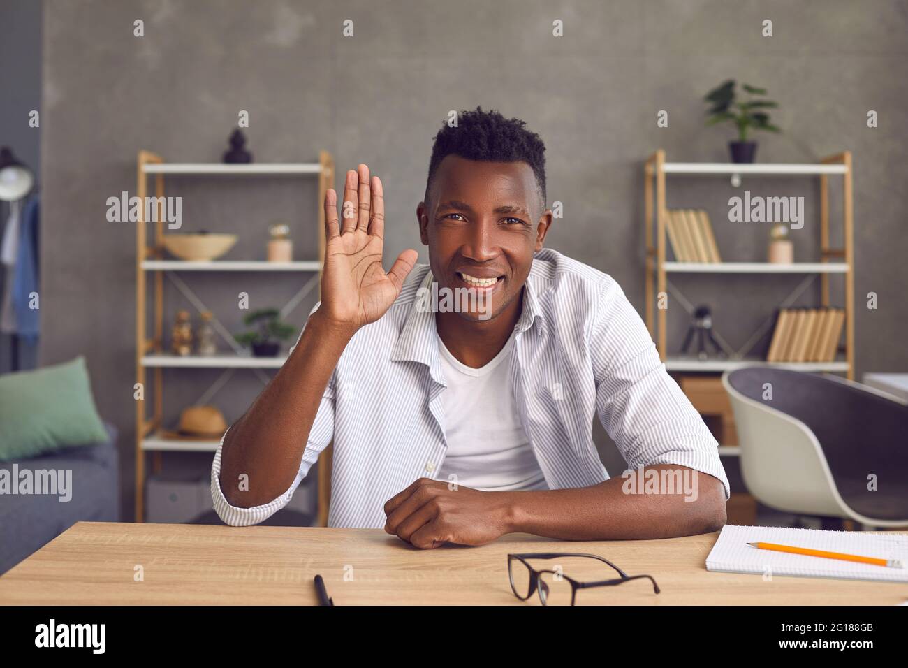 Felice studente nero seduto alla sua scrivania a casa, sorridente e sventolante mano alla macchina fotografica Foto Stock