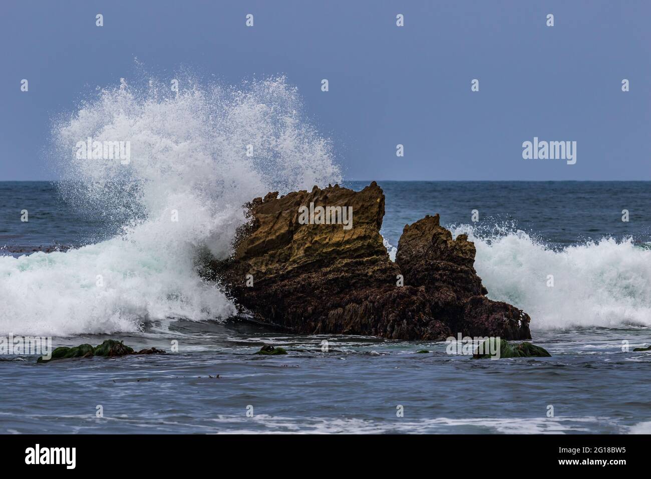 L'onda dell'oceano che si infrangono sulla roccia appena offshore a Laguna Beach, California. Spray bianco nell'aria; cielo blu sullo sfondo. Foto Stock
