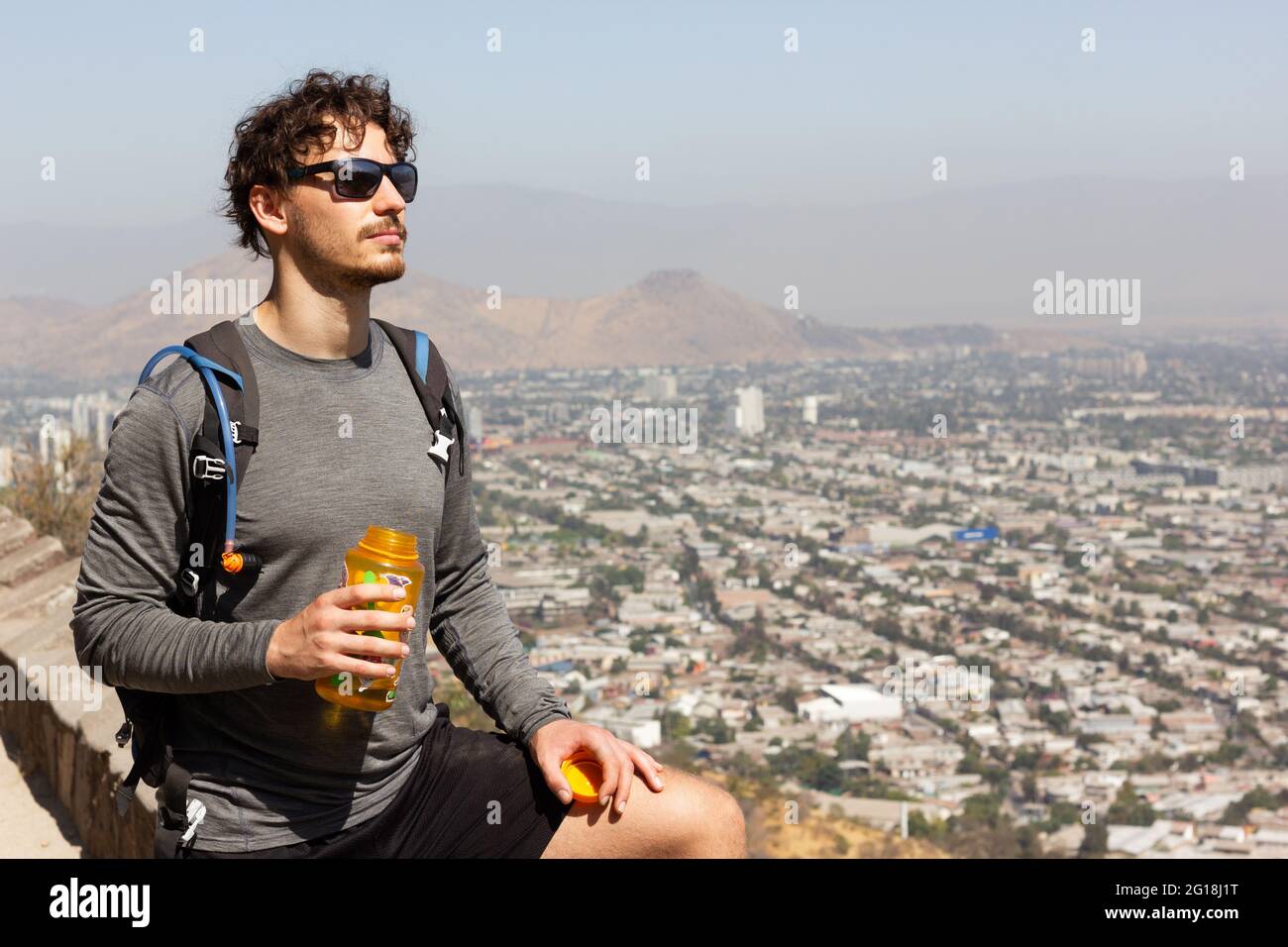 Giovane uomo con occhiali da sole che tengono una bottiglia d'acqua e che  gode di splendide vedute della città di Santiago in Cile durante il giorno  di sole. Escursionista facendo una pausa