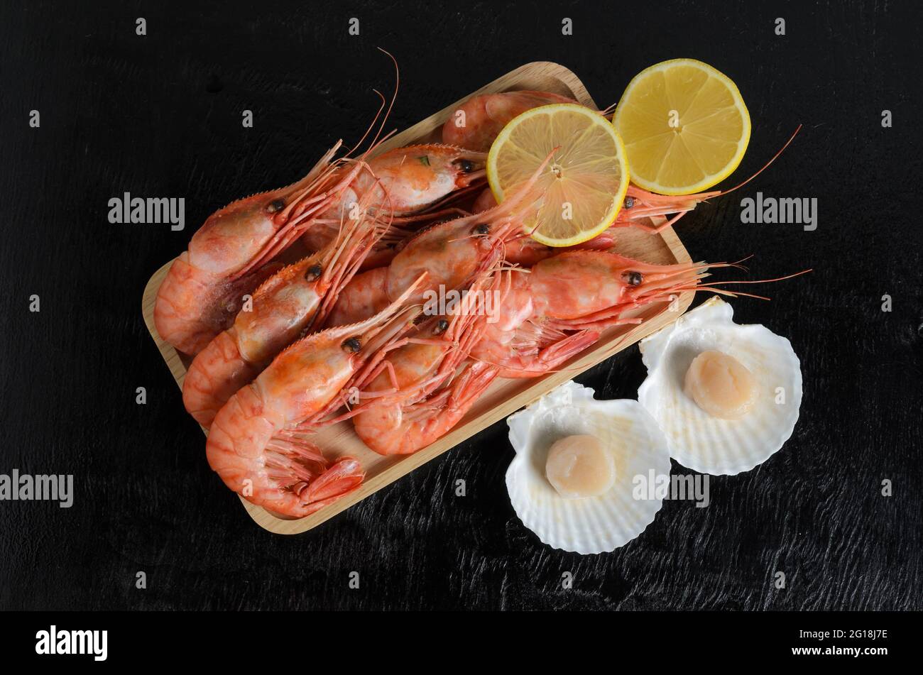 I gamberetti bolliti in una conchiglia con limone in una ciotola di legno con un capesante crudo in una conchiglia su uno sfondo di legno scuro. Messa a fuoco selettiva. Foto Stock