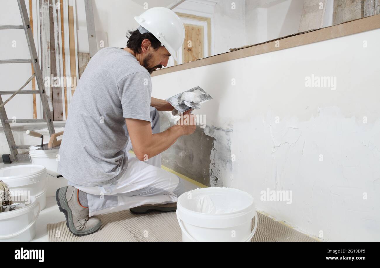 uomo addetto alla costruzione di intonaci al lavoro, prende gesso da secchio e lo mette su cazzuola per intonacare la parete, indossa casco all'interno del palazzo si Foto Stock