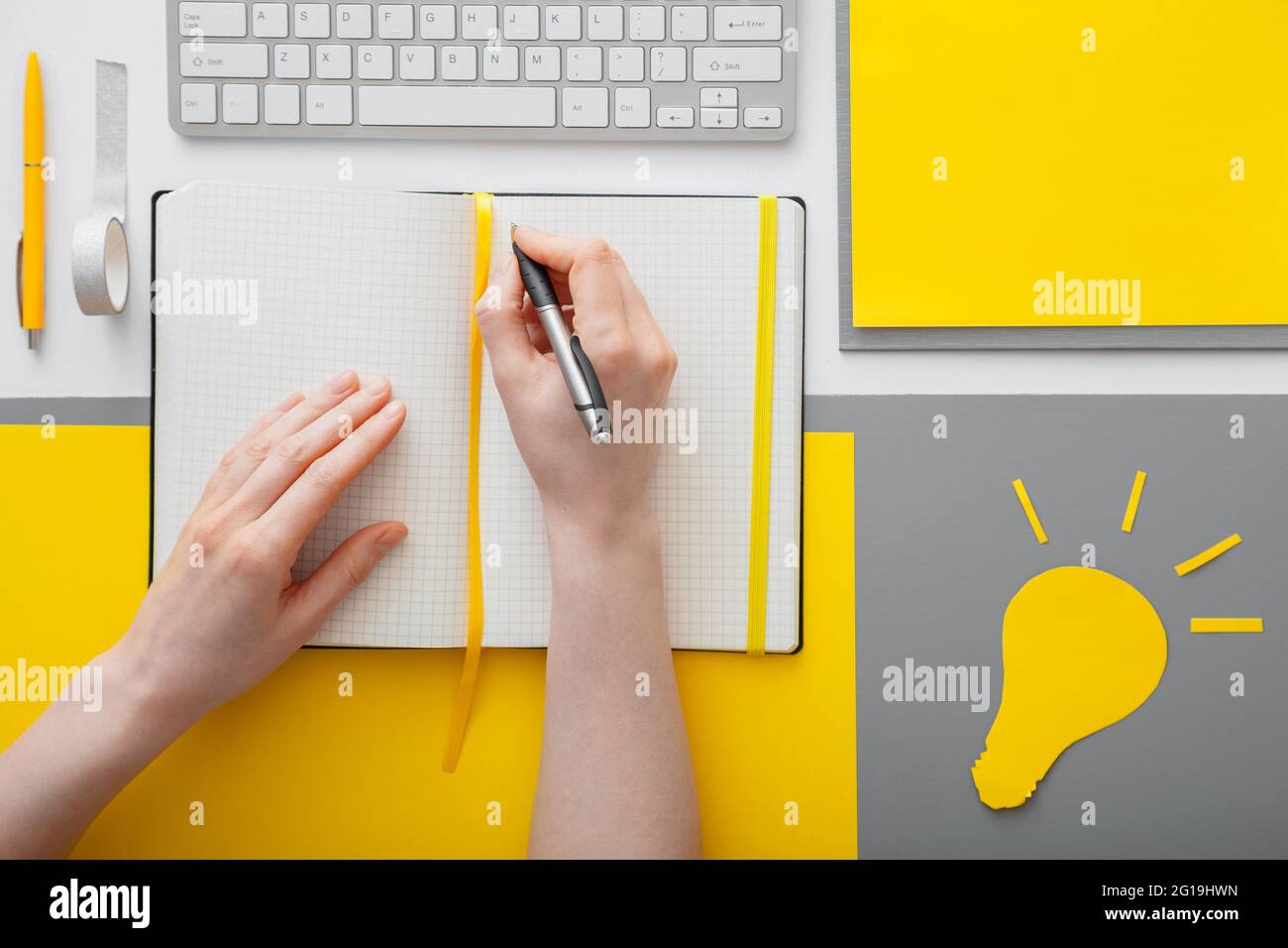 Le mani femminili scrivono un obiettivo di idea in un notebook vuoto sul desktop nello spazio di lavoro. La donna scrive l'elenco delle idee in taccuino su sfondo grigio giallo Foto Stock