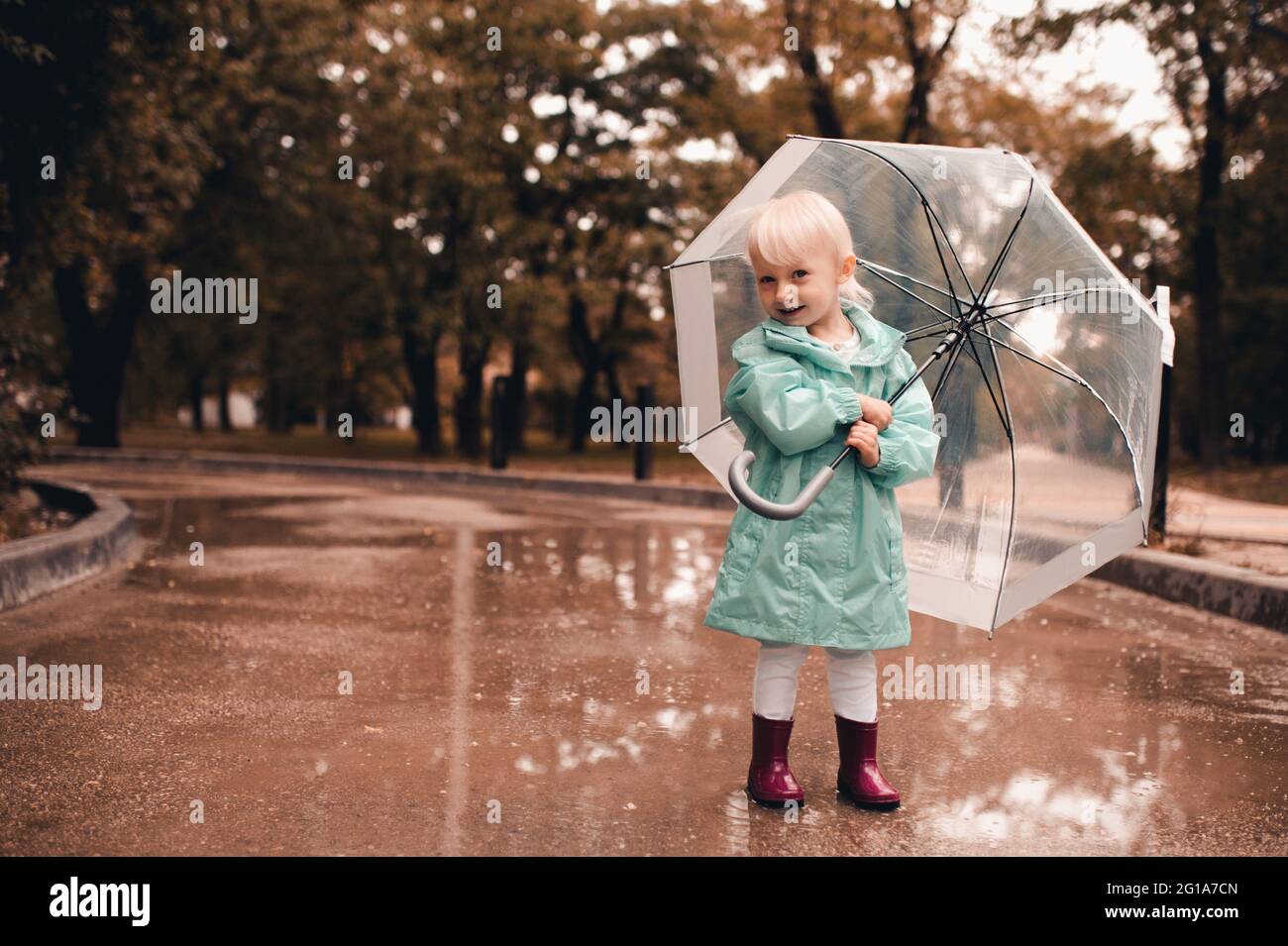 Ragazza divertente bambino 3-4 anni che tiene ombrello indossare arcobaleno e impermeabile nel parco camminare in pozzanghere all'aperto. Guardando la fotocamera. Stagione autunnale. Bambino Foto Stock
