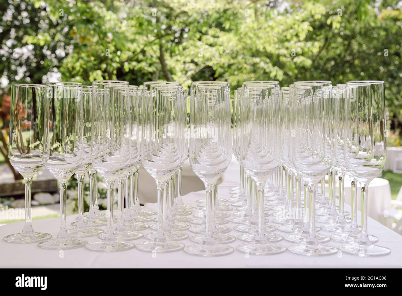 Decine di bicchieri da vino su un tavolo pronti per essere utilizzati per una festa o una festa, per un servizio di catering per matrimoni, con alberi verdi sullo sfondo Foto Stock