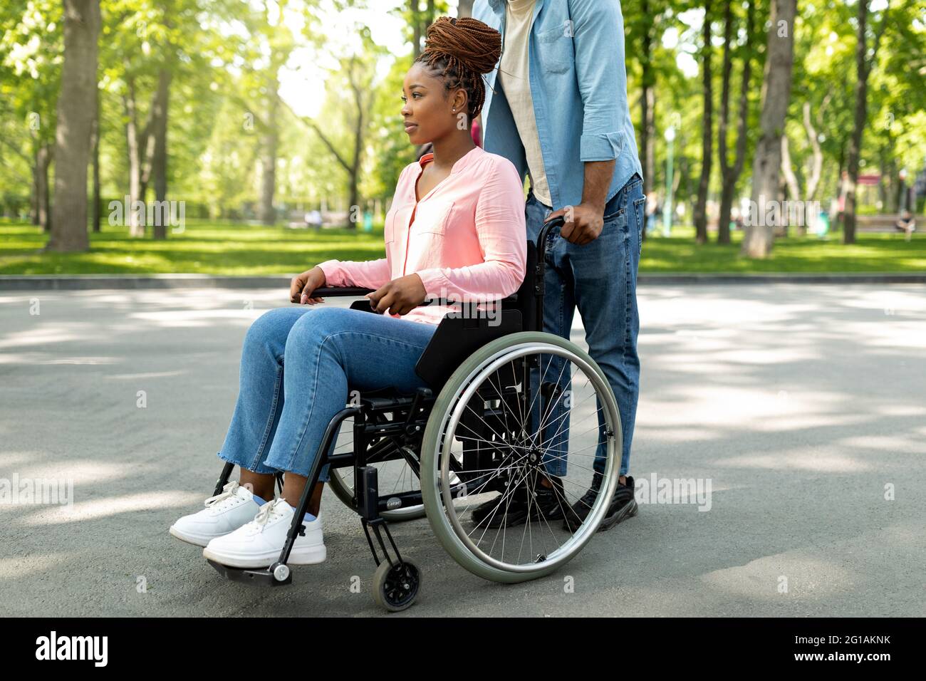 Donna nera disabile in sedia a rotelle a piedi con il suo affettuoso marito all'aperto, trascorrendo molto tempo insieme Foto Stock