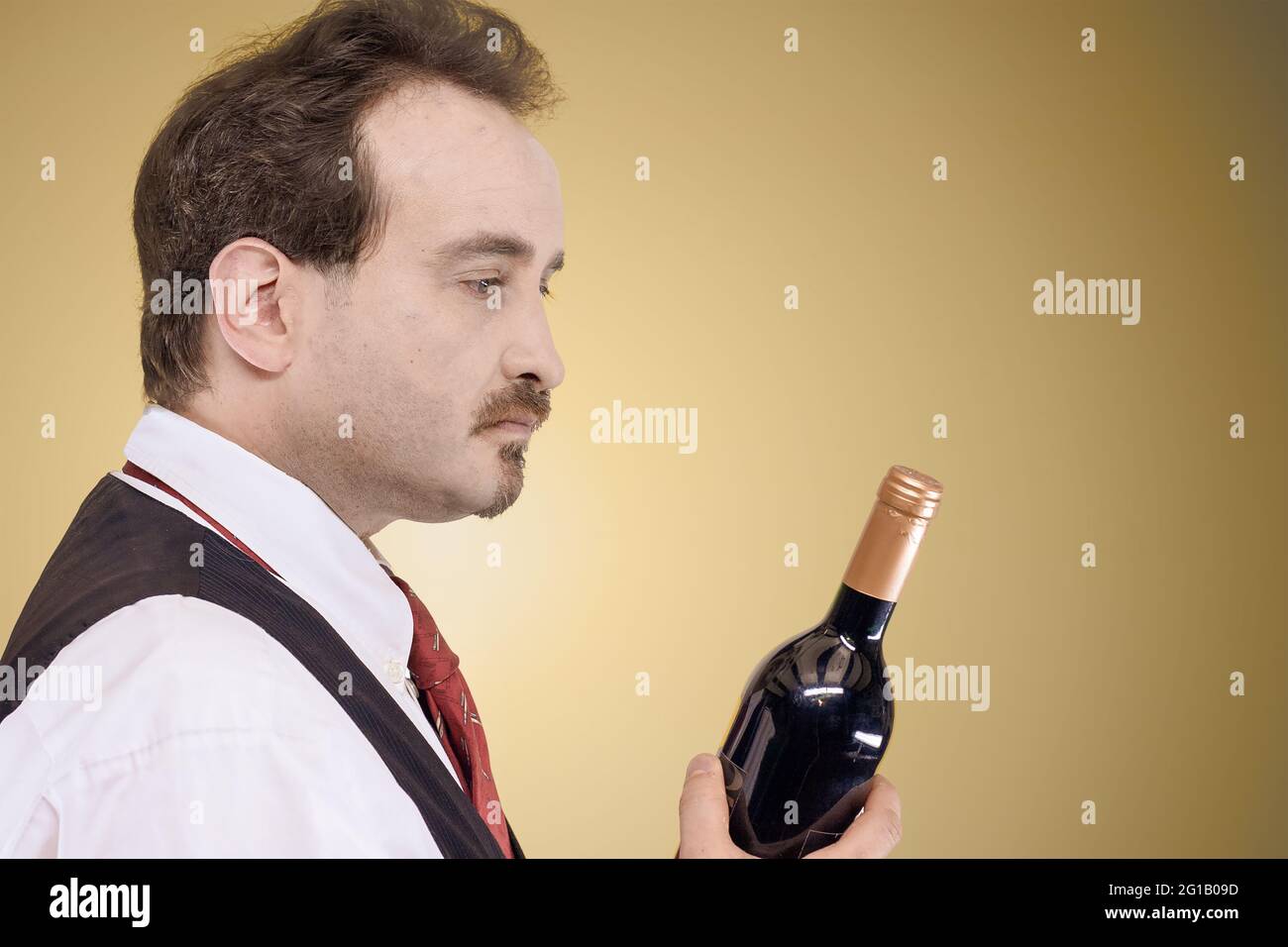 Uomo in camicia bianca, gilet e cravatta guardando una bottiglia di vino davanti a uno sfondo pastello chiaro Foto Stock