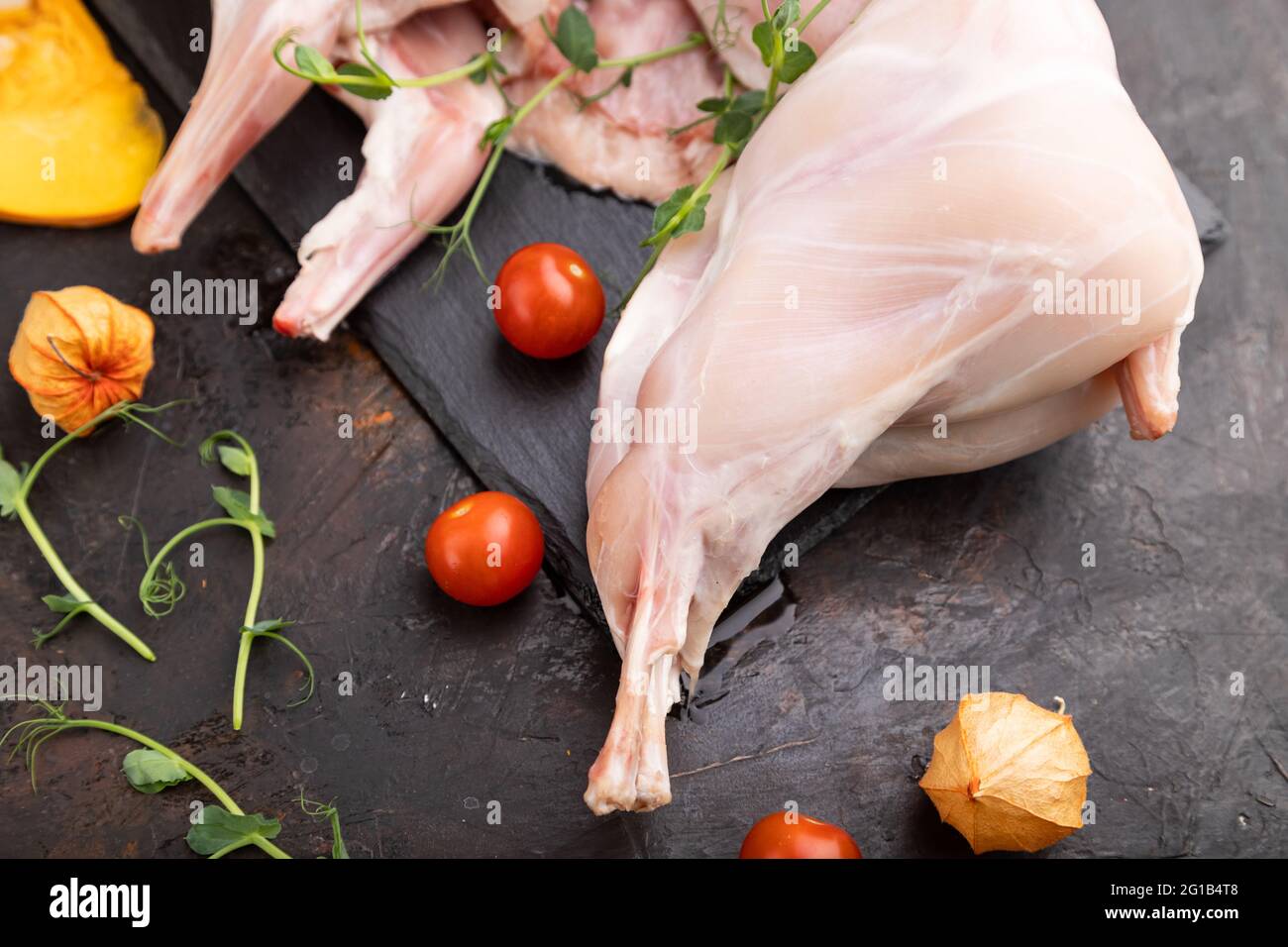 Coniglio intero crudo con zucca, pomodori, germogli di piselli su fondo nero di cemento e tessuto di lino. Vista laterale, primo piano, messa a fuoco selettiva. Foto Stock