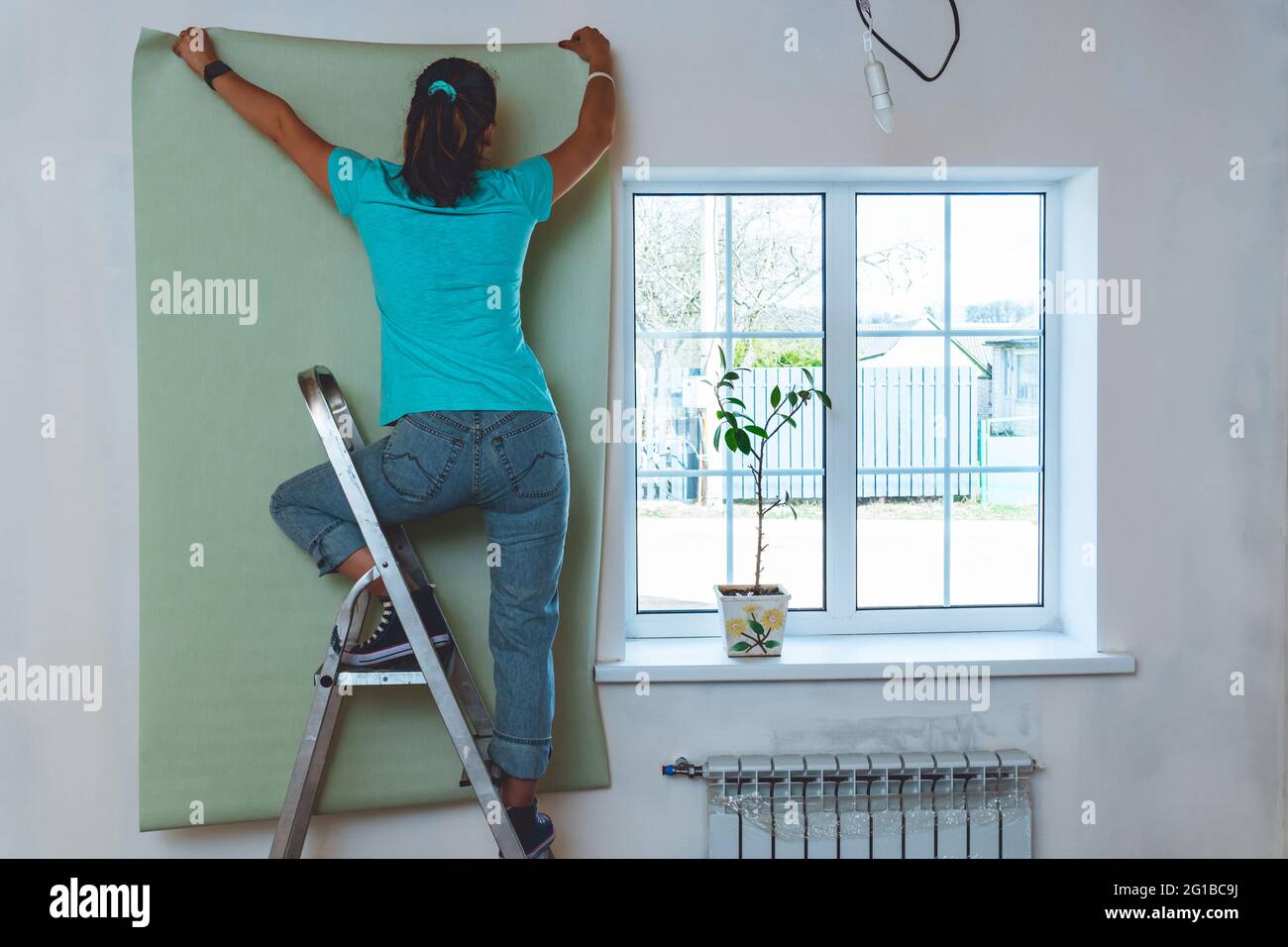 Donna in jeans e T-shirt turchese in piedi sulla scala e tenendo un pezzo di carta da parati. Ristrutturazione e miglioramento della casa. Foto Stock