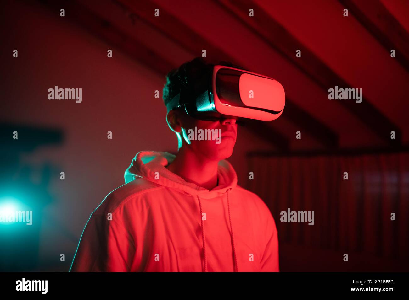 Uomo che indossa una felpa con cappuccio mentre usa occhiali VR e in piedi in studio con luci al neon rosse Foto Stock