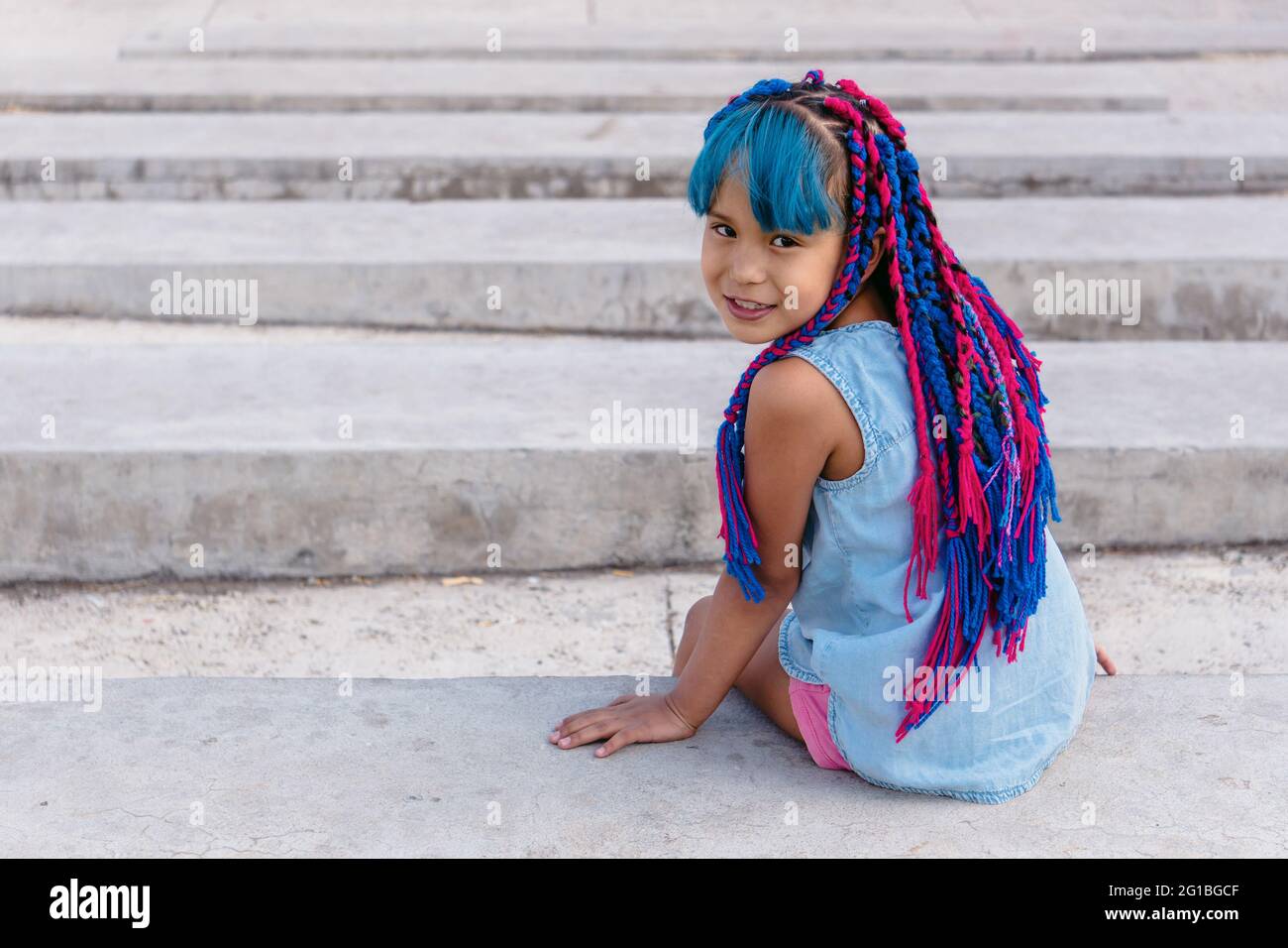 Allegro bambino etnico carino con trecce colorate seduti su scale di cemento mentre si guarda la fotocamera in luce del giorno Foto Stock