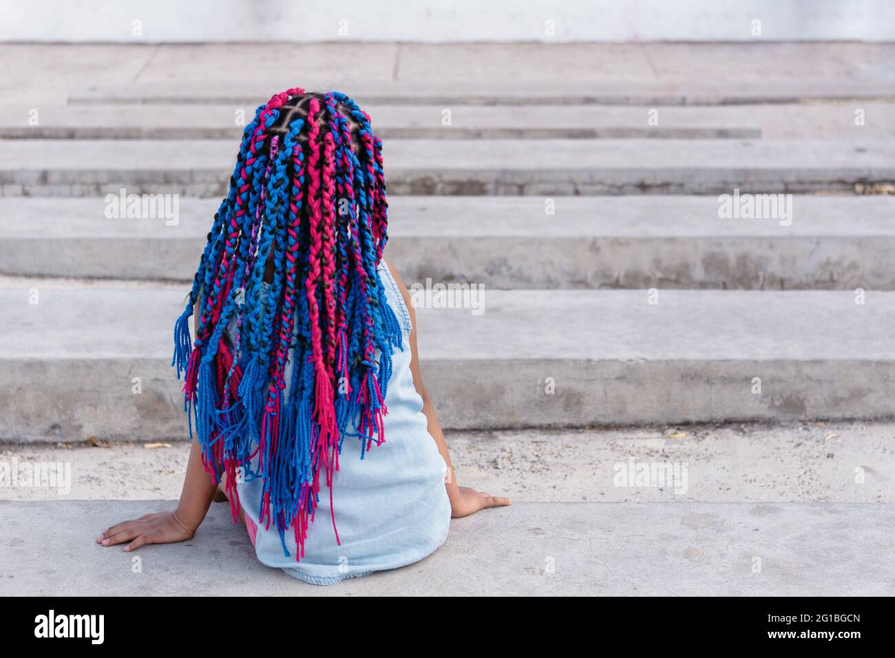 Vista sul retro di un bambino etnico irriconoscibile e sognante, con trecce colorate che si trovano su una scala in cemento e che si affaccia alla luce del giorno Foto Stock