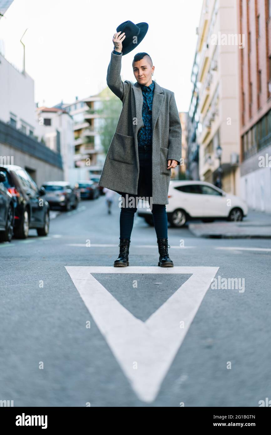 Persona transgender in elegante cappotto e cappello in piedi con braccio sollevato sulla strada urbana mentre si guarda la fotocamera Foto Stock