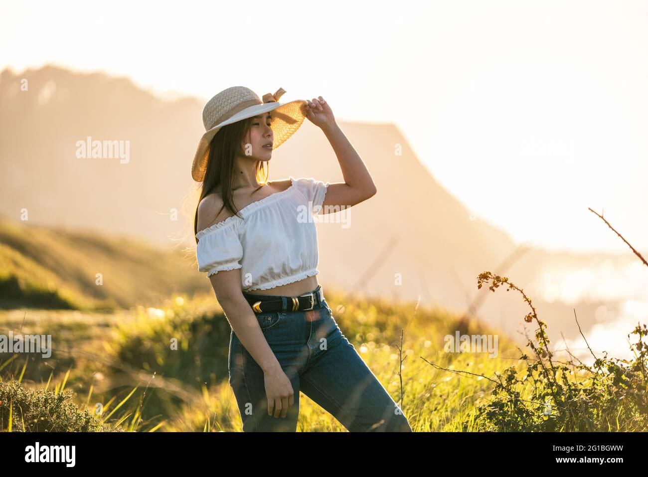 Attraente donna sognante in jeans e cappello bianco che tocca la cima brim e guardando via in pensieri piacevoli mentre si levano in piedi su terreno erboso lussureggiante sul sole Foto Stock