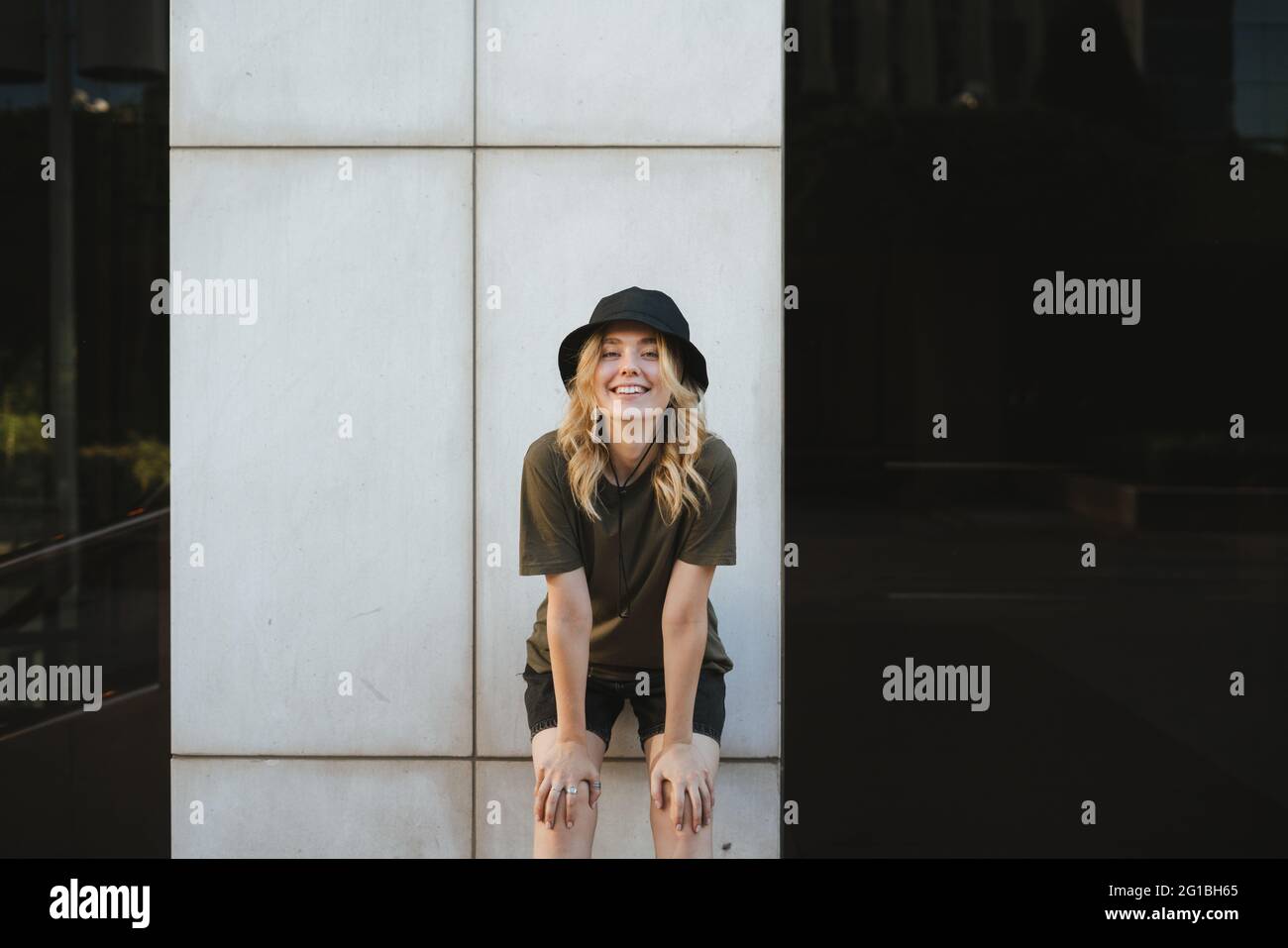 Sorridente giovane donna in un abbigliamento casual che si inclina in avanti mentre si guarda la fotocamera in città alla luce del giorno Foto Stock