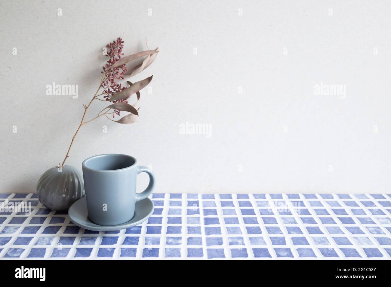 Tazzina da caffè grigia e vaso di fiori secchi su tavola di tegole in ceramica blu. Fondo parete bianco. Interni domestici Foto Stock