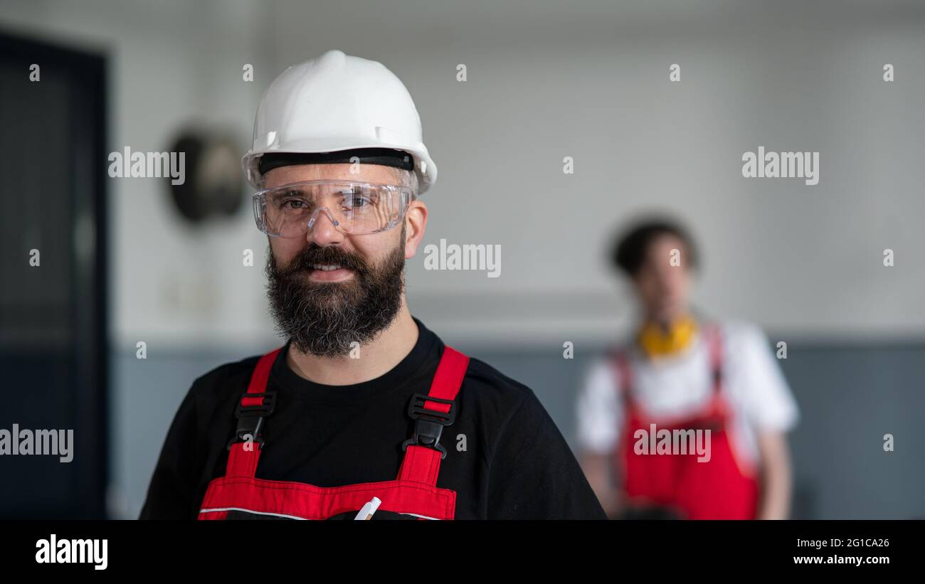 Ritratto del lavoratore con casco e occhiali protettivi in interni in fabbrica guardando la fotocamera. Foto Stock