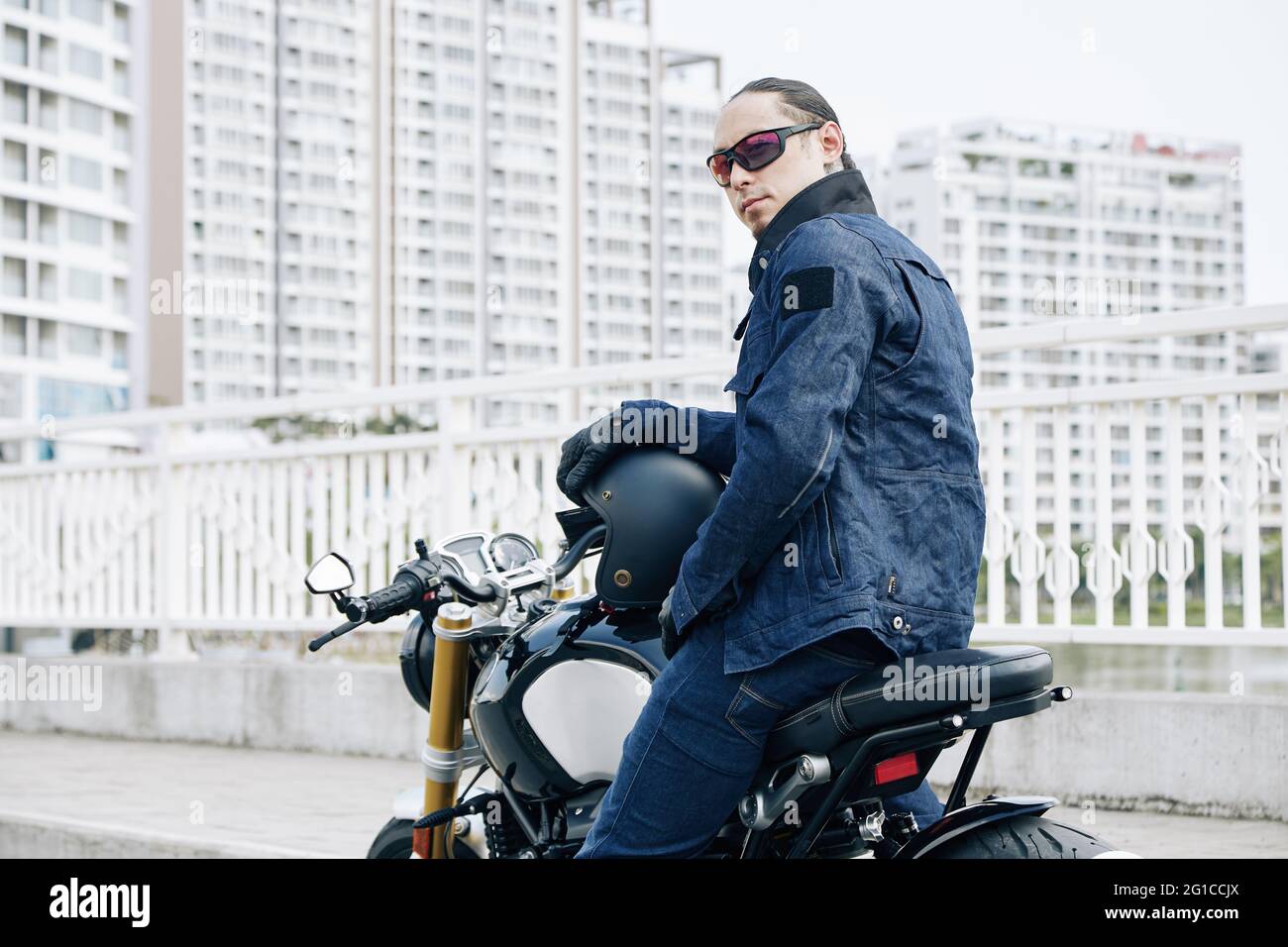 Uomo serio con giacca in denim e occhiali da sole seduti sulla moto e tornando alla macchina fotografica Foto Stock