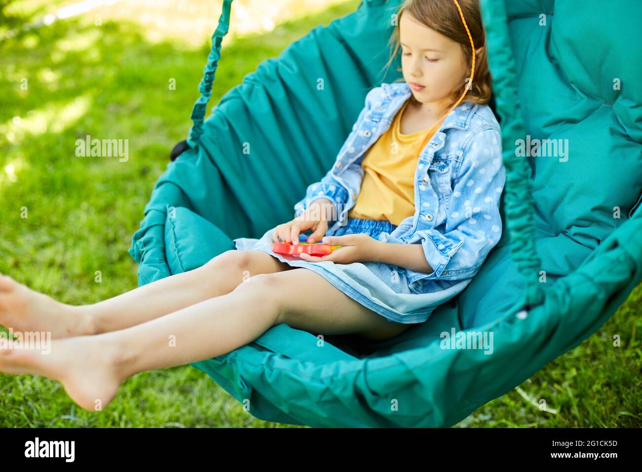 Una bambina la sedia appesa all'aperto gioco pop it, mani di bambino che giocano bolle di un giocattolo iridato destrecessing, giocattolo fidget nel cortile di hou Foto Stock