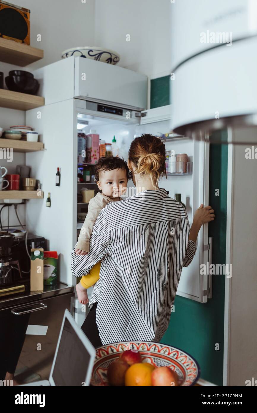 Vista posteriore della madre che trasporta il bambino mentre apre il frigorifero in cucina Foto Stock