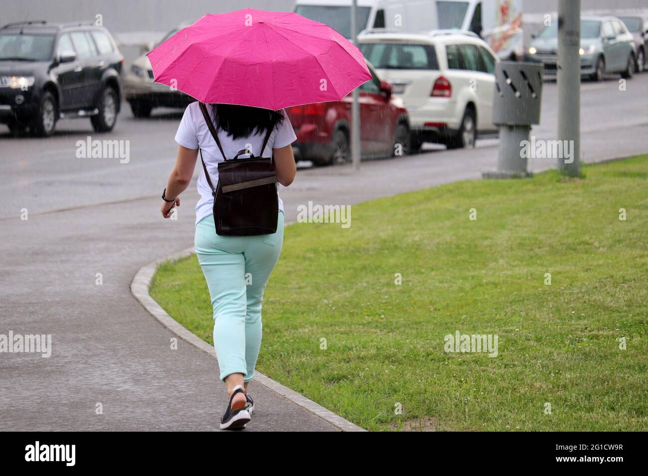 Pioggia in città, donna con ombrello rosa che cammina su una strada sullo sfondo di un ingorgo. Automobili che guidano su una strada in tempo piovoso, tempesta estiva Foto Stock