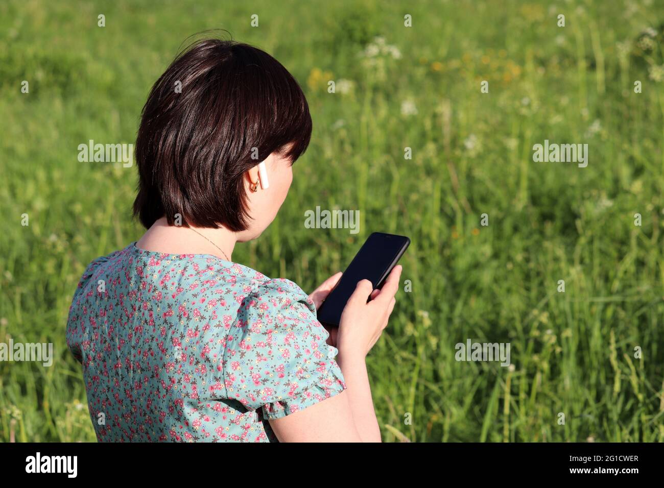 Donna in cuffia wireless che utilizza lo smartphone su un prato estivo. Mani femminili con telefono cellulare su sfondo verde natura Foto Stock
