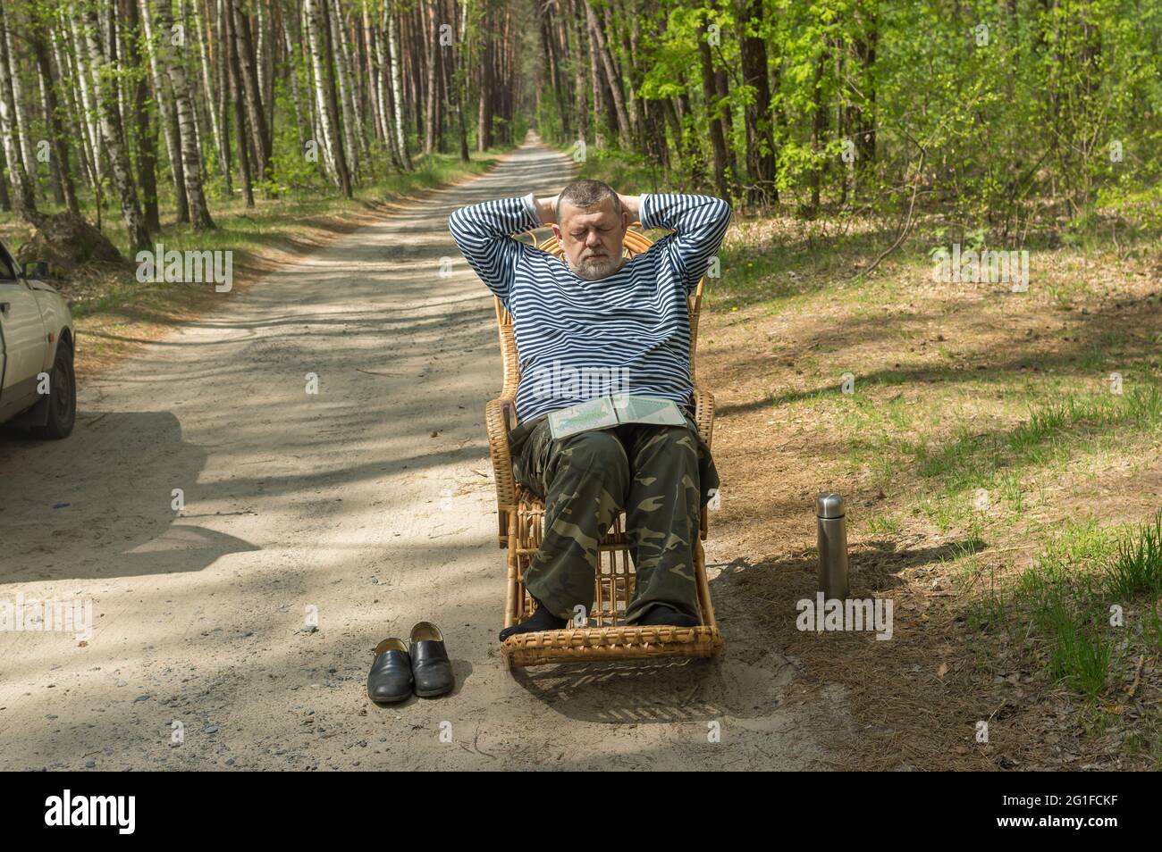 Autista caucasico senior che riposa fuori della sua auto dormendo in una sedia a dondolo in vimini nella foresta primaverile Foto Stock