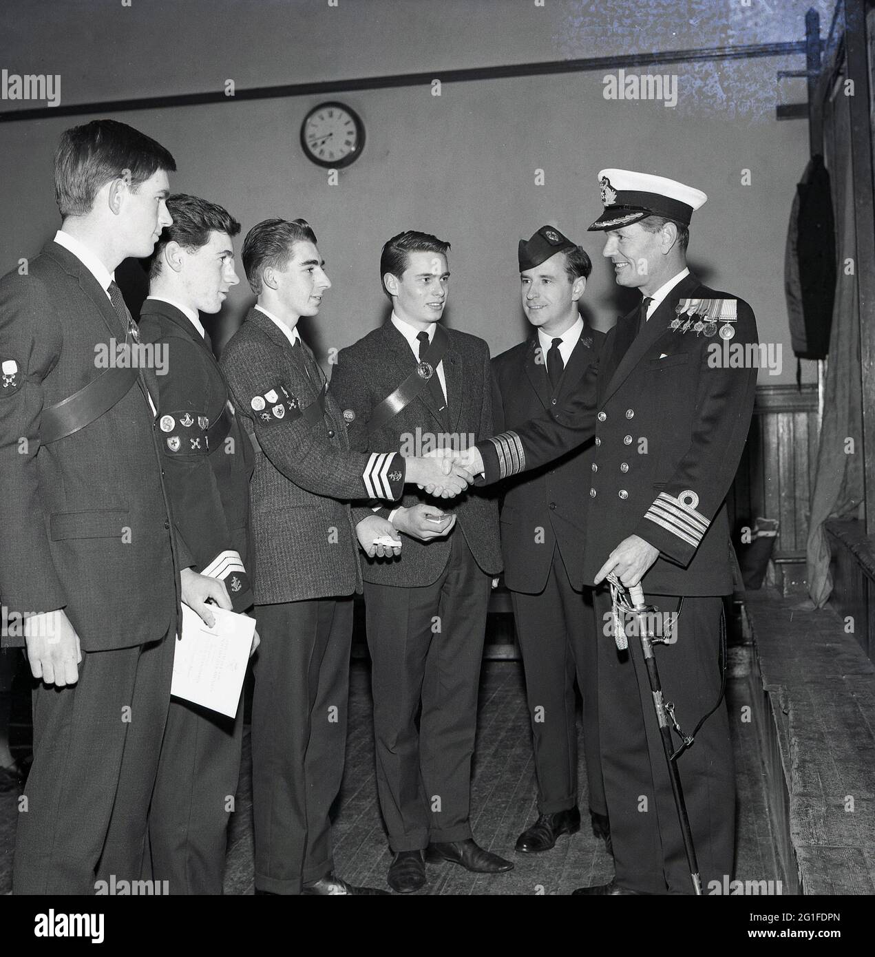 Anni '60, storico, congratulazioni .... un capitano Royal Naval decorato con la spada, scuotendo le mani con un giovane orgoglioso, in piedi insieme con altri tre membri maschi della Brigata Boys in una cerimonia di premiazione, Fife, Scozia, UK. Fondata a Glasgow, in Scozia, da William Alexander Smith nel 1883, per combinare perforazione e divertimento con valori cristiani, la Brigata dei ragazzi è la più grande organizzazione giovanile cristiana in uniforme del Regno Unito. L'introduzione del campeggio per adolescenti da parte di Alexander Smith fu qualcosa di nuovo alla fine del XIX secolo, con il primo campo BB a Tighnabruiach nel 1886. Foto Stock