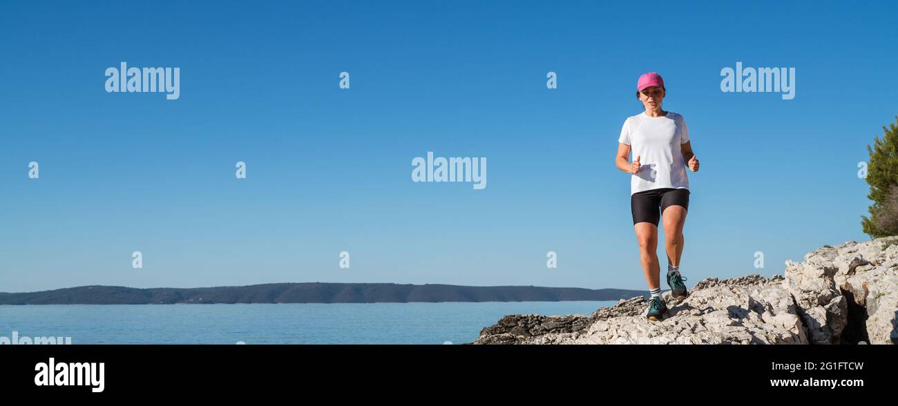 Giovane donna vestito da running abiti sportivi e berretto rosa godendo di jogging al mattino vicino alla costa rocciosa e tranquilla del mare. Le persone sportive trainano attività di corsa Foto Stock