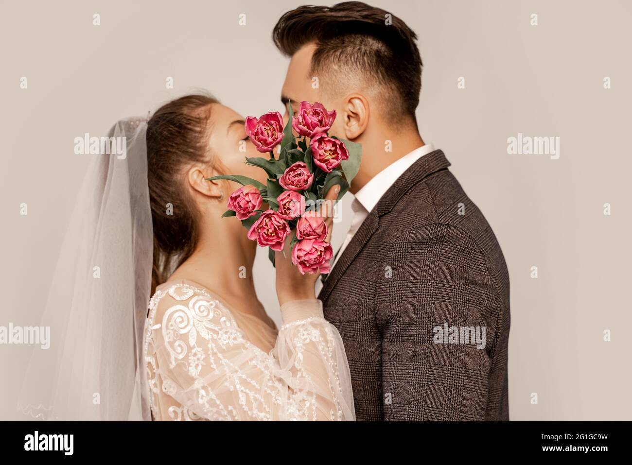 giovani sposi che baciano mentre oscurano i volti con tulipani isolati in grigio Foto Stock