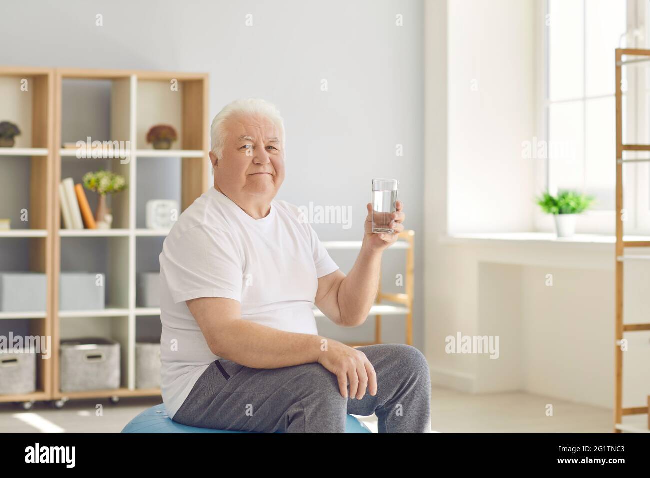 Uomo anziano che tiene un bicchiere d'acqua seduto su un flipper a casa e guardando la fotocamera. Foto Stock