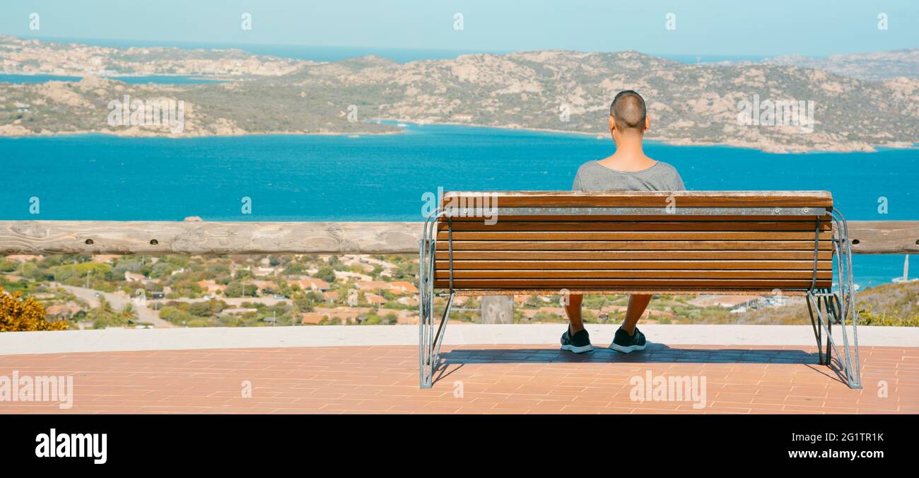 Giovane caucasico seduto in una panchina a Palau, sulla costa settentrionale della Sardegna, Italia, osserva il Mar Mediterraneo e l'Arcipelago della Maddalena Foto Stock