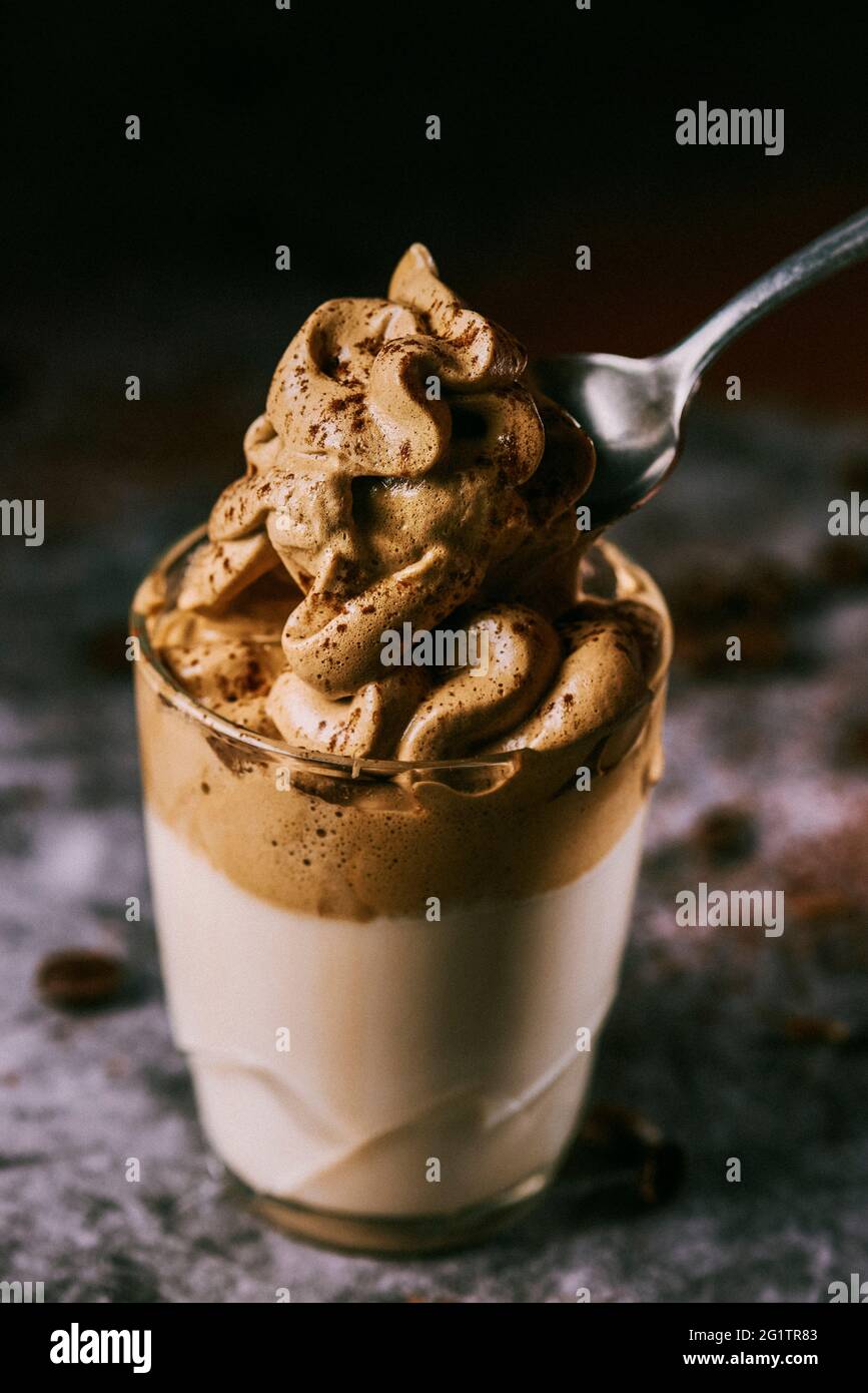 qualcuno prende un cucchiaio di caffè dalgona da un bicchiere, su una superficie di pietra grigia scura contro uno sfondo nero Foto Stock