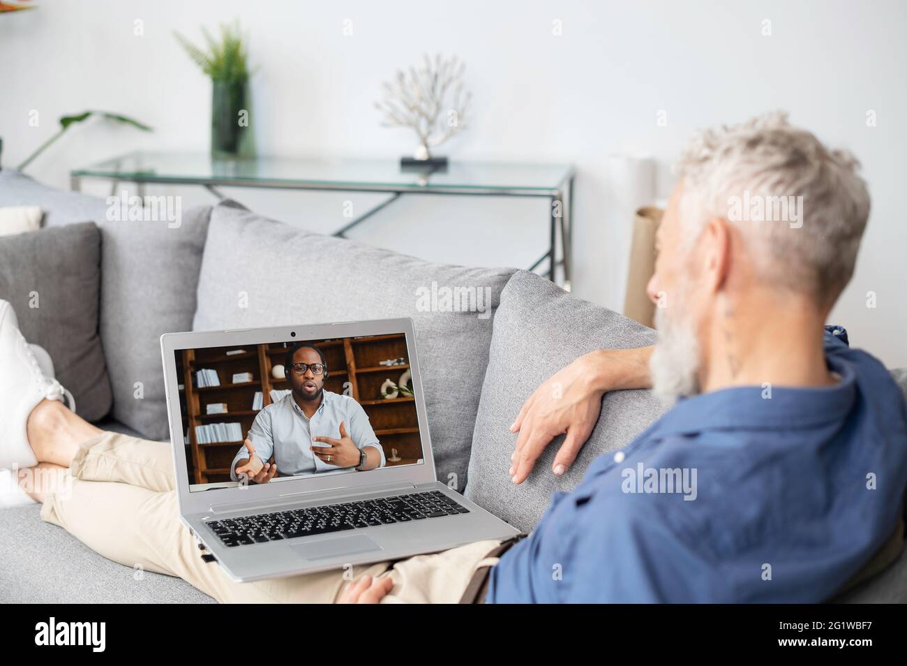 Vista posteriore sulla spalla di un uomo maturo sullo schermo del laptop con un uomo afroamericano su di esso, due diversi colleghi di sesso maschile che parlano online, utilizzando l'app per computer per la connessione video Foto Stock