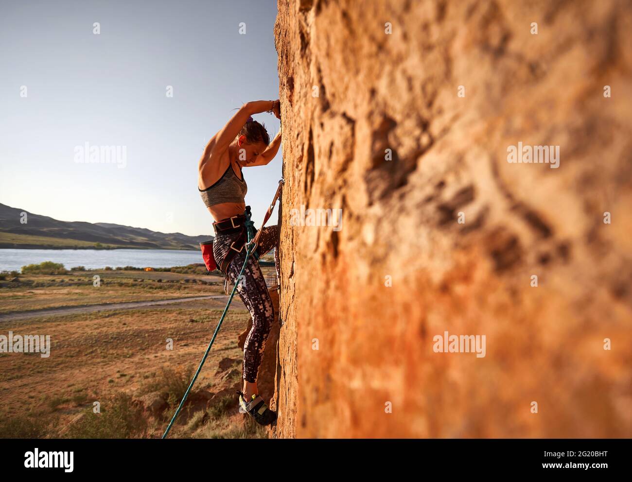 La giovane atleta sta arrampicando sulla parete piatta alta della roccia al tramonto vicino al fiume in Kazakhstan Foto Stock