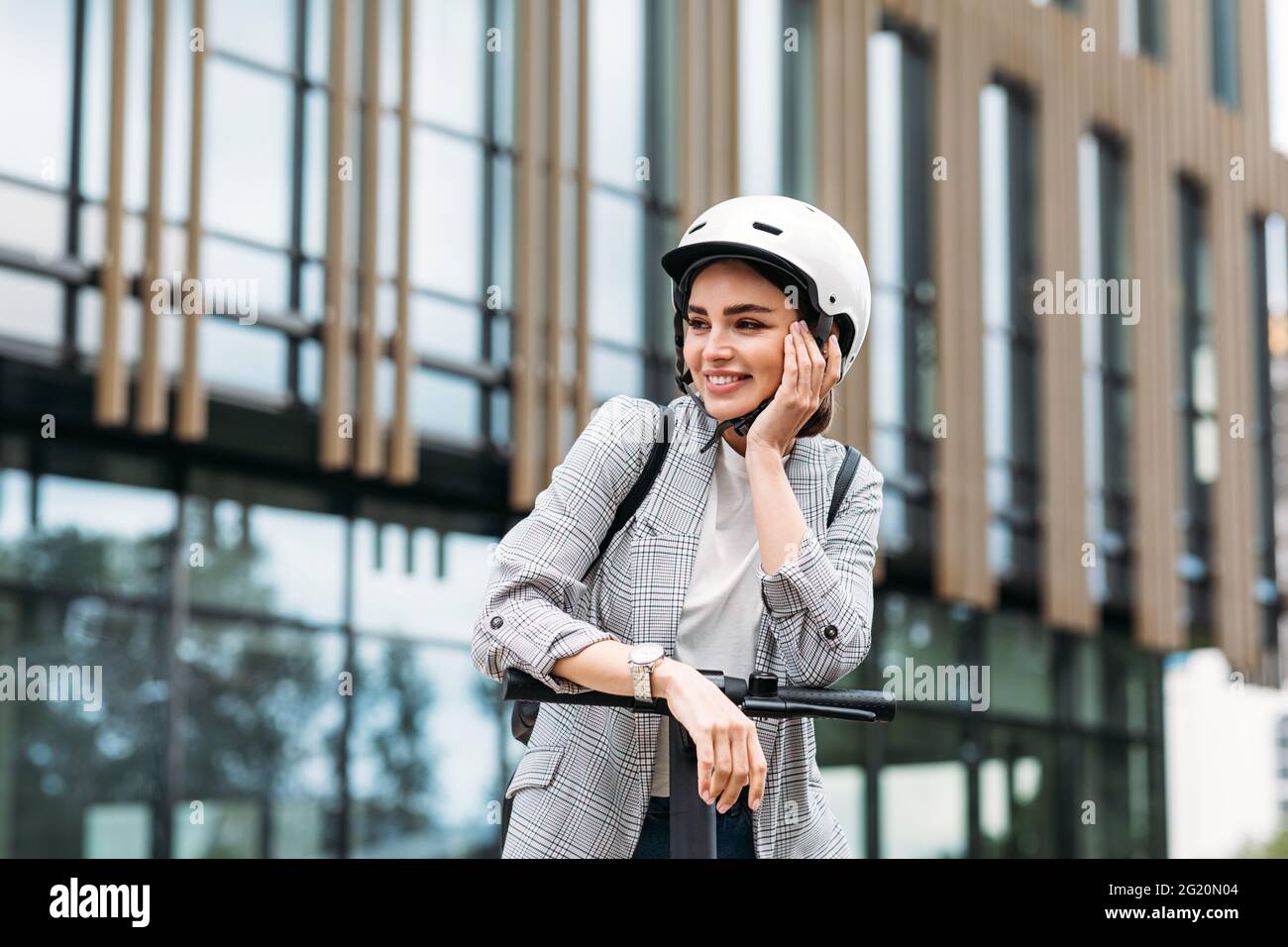 Donna positiva appoggiata sul manubrio dello scooter elettrico a spinta. Bella donna in casco guardando lontano mentre si trova in città. Foto Stock
