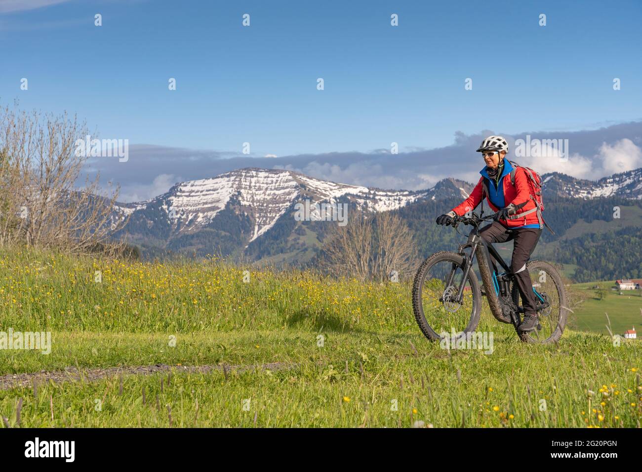 Donna anziana sorridente che guida la sua mountain bike elettrica sotto le montagne innevate della catena Nagelfluh vicino Oberstaufen, Allgaeu, Baviera, Germania Foto Stock