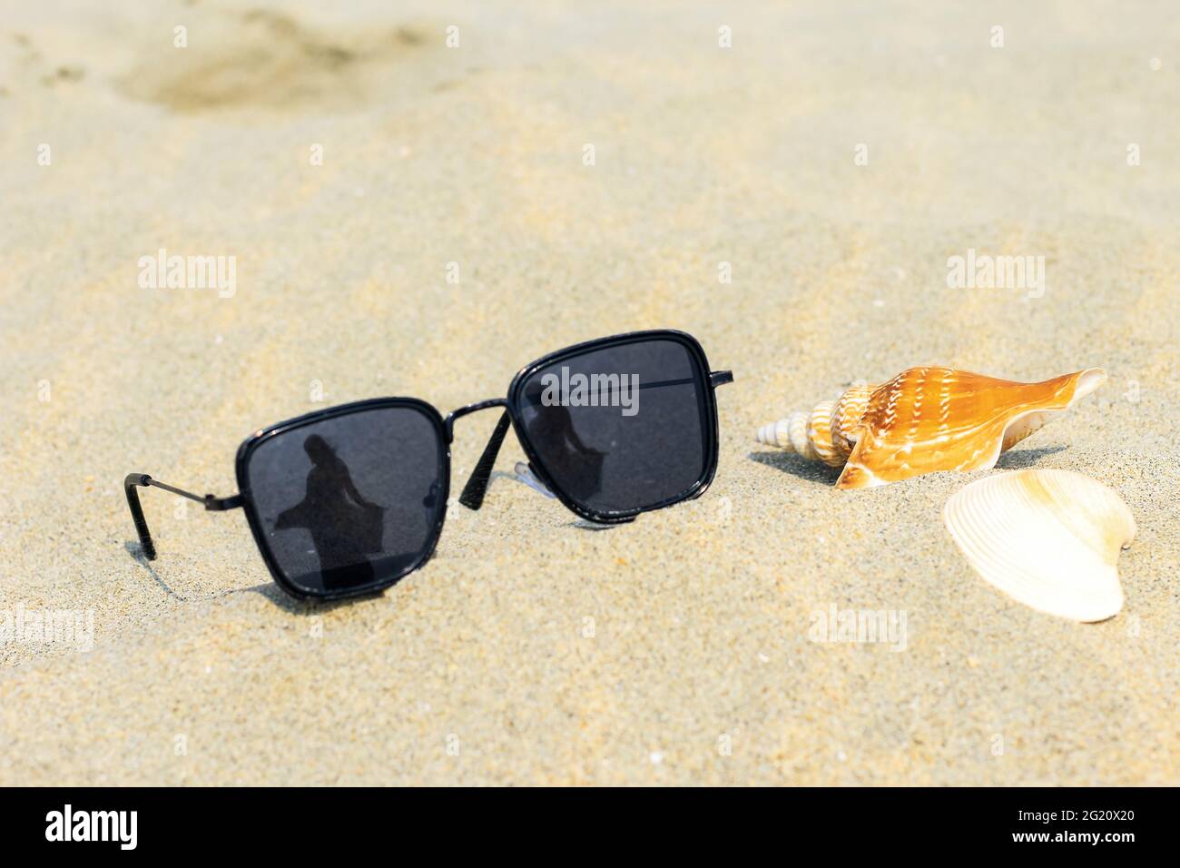 Occhiali da sole riflettenti su una spiaggia di sabbia in estate, e il  cielo blu con una ragazza si riflette in occhiali con conchiglie. Il  riflesso della ragazza sul vetro Foto stock -