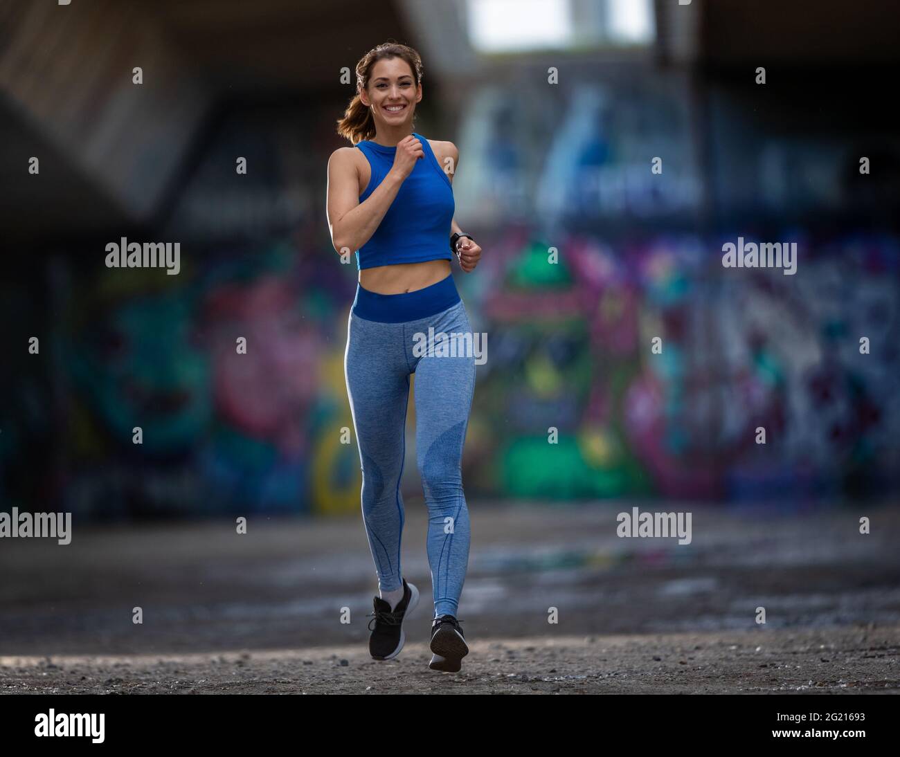 Fit giovane donna jogging in zona urbana. L'atleta cammina a velocità sorridente con la parete coperta di graffiti sullo sfondo. Foto Stock