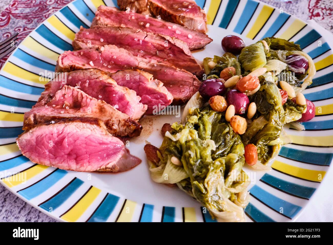 Un piatto di bistecca di ferro piatto cucinato raro con insalata di escarole cotte, olive, uvetta e pinoli come contorno. La bistecca di ferro piatta è molto morbida Foto Stock