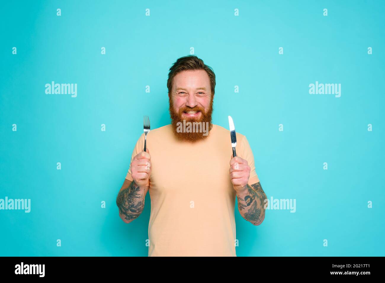 L'uomo felice con i tatuaggi è pronto a mangiare con le posate in mano Foto Stock