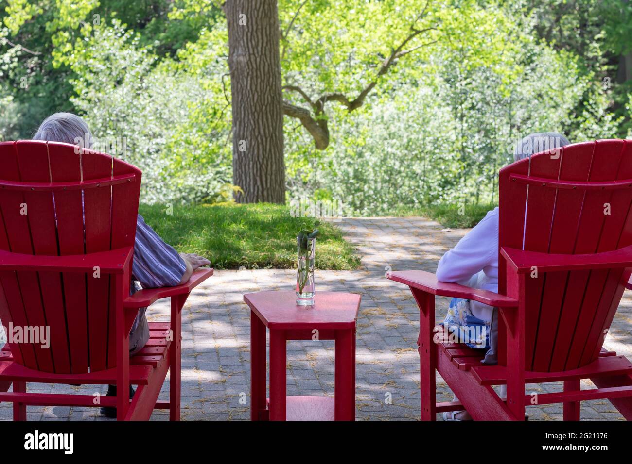 Vista posteriore della coppia più anziana - uomo e donna - negli anni '70, seduti in rosso Muskoka o Adirondack sedie sul cortile patio con vista sugli alberi in estate. Foto Stock