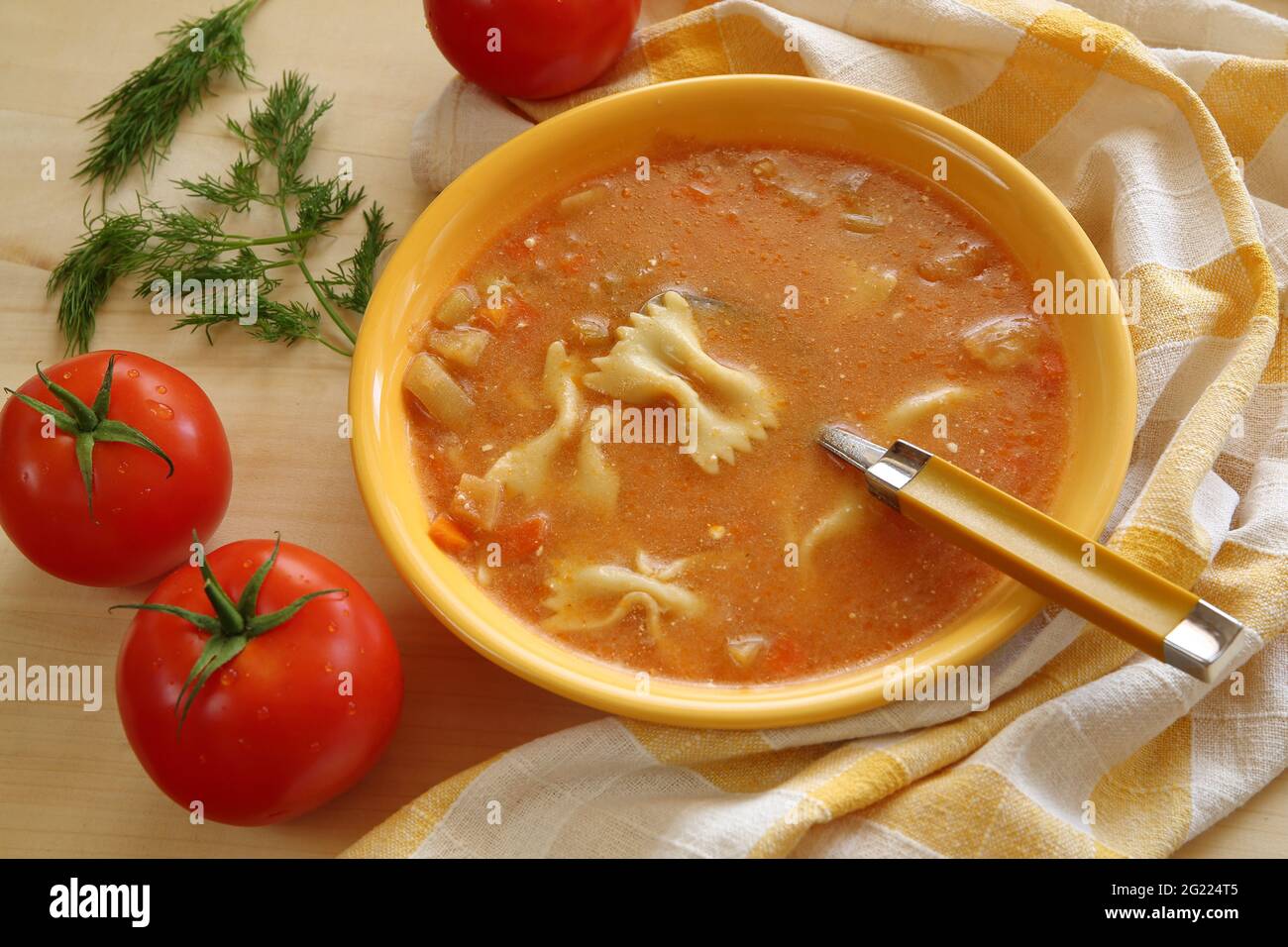 Tradizionale zuppa di pomodoro fatta in casa in ciotola gialla, con nuddles e cucchiaio, tre pomodori rossi maturi, erbe verdi, biancheria da cucina. Foto Stock